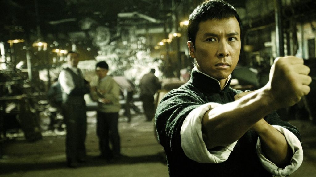 A martial artist stands in a Wing Chun fighting stance inside a dimly lit workshop, extending one arm forward while focusing on an unseen opponent in a scene from Ip Man.