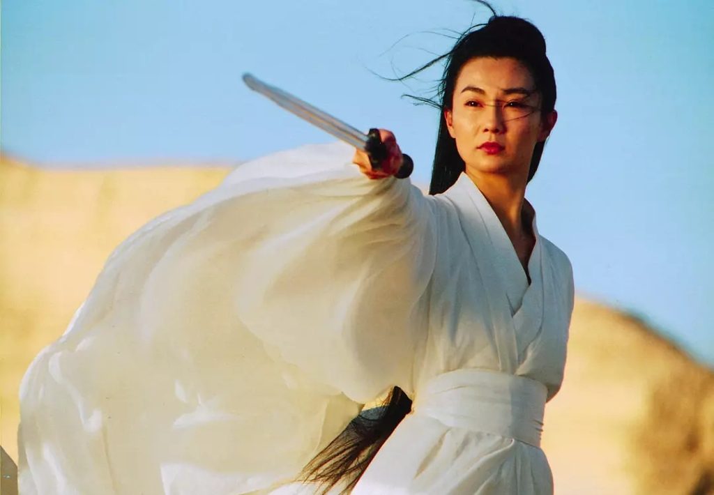 Maggie Cheung, dressed in flowing white robes, points a sword forward in a windswept desert landscape in a scene from Hero (2002).
