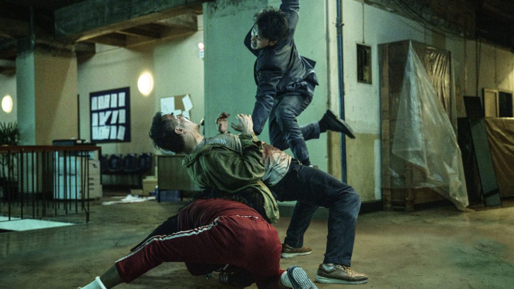 A fighter in a dark suit leaps through the air while attacking multiple opponents during a frenetic hand-to-hand combat sequence inside a dimly lit building in a scene from The Furious.