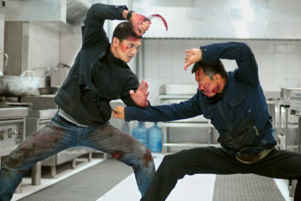 Two bloodied fighters clash with knives inside a commercial kitchen, each bracing for a lethal strike during an intense hand-to-hand combat sequence in The Raid 2.