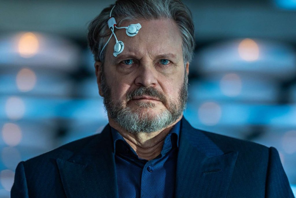 Close-up portrait of a middle-aged man in a dark suit with a medical monitoring device attached to his temple, staring intently ahead in a blue-lit, high-tech environment.