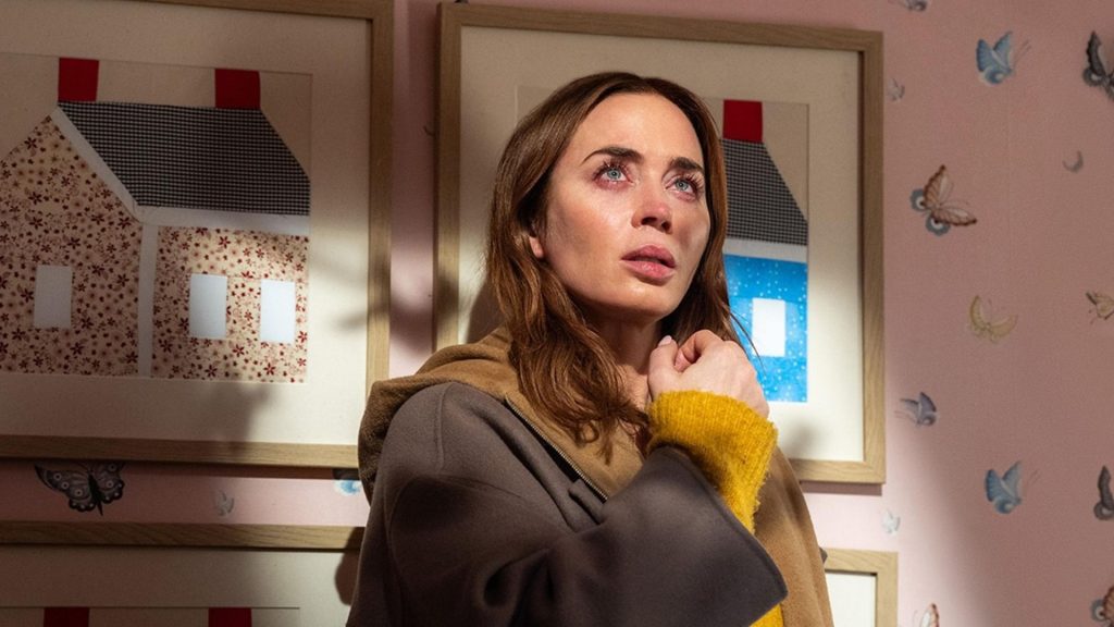 Emily Blunt as Margaret Fairchild in Disclosure Day, standing against a pink wallpapered wall with framed house illustrations, her expression stricken and searching.