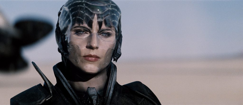 Close-up of a futuristic armored warrior wearing a transparent biomechanical helmet and dark combat suit, staring ahead with a stern expression against a muted sky backdrop.