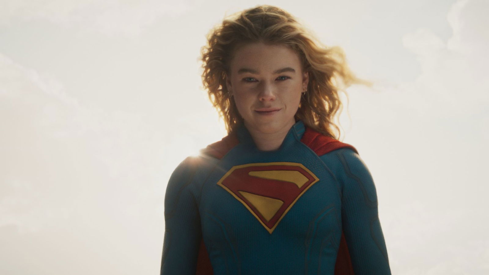 A superhero in a blue suit and red cape (supergirl) looks downward with a calm expression while standing against a bright, sunlit sky. She is one of the strongest kryptonians

