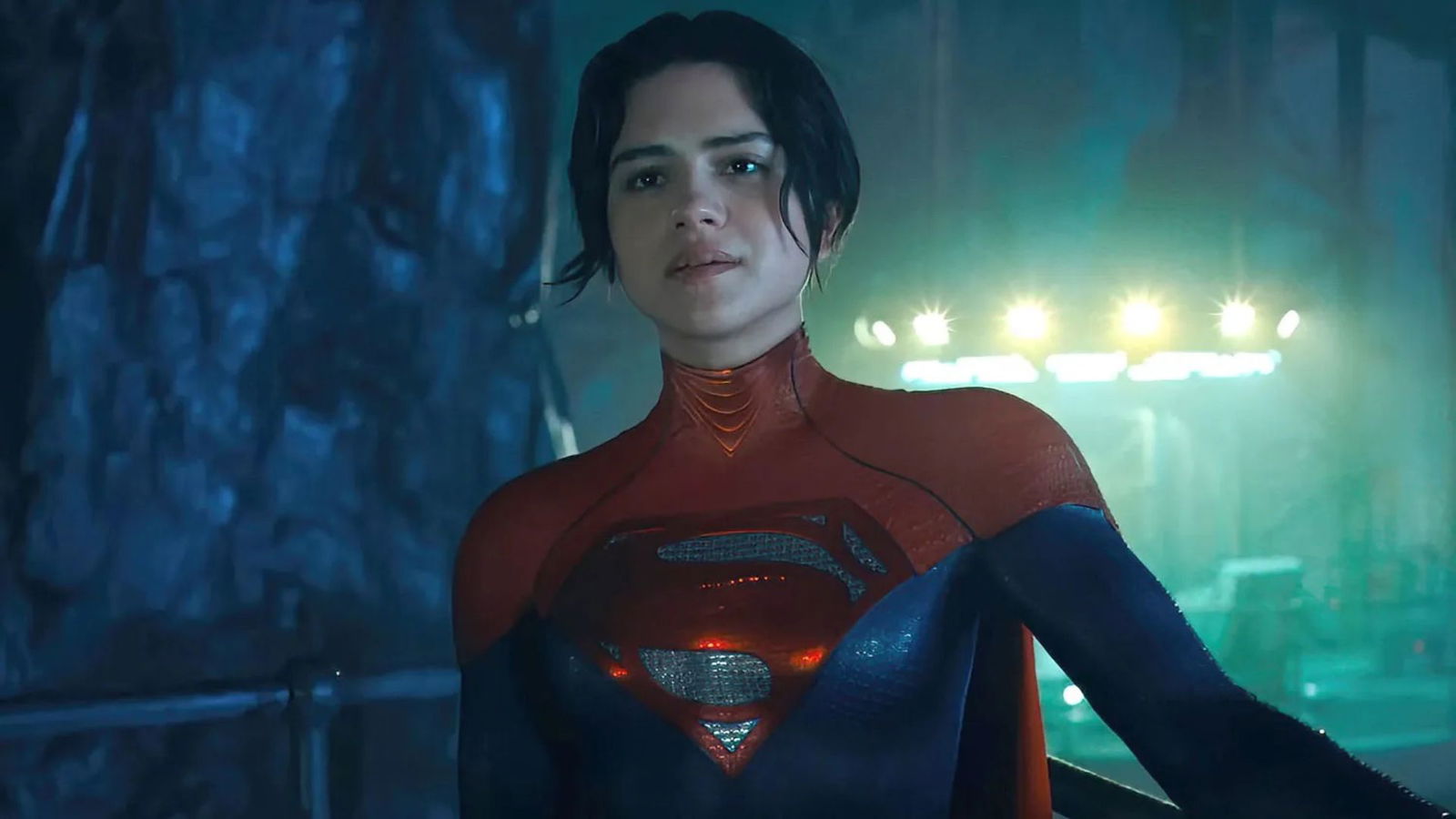 A superhero wearing a darker red-and-blue suit stands in a dimly lit industrial setting, illuminated by bright lights in the background.