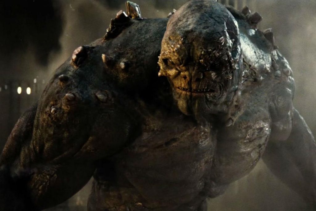 A massive gray-skinned creature covered in bony protrusions and jagged spikes looms forward through smoke and debris, displaying immense strength and an intimidating presence.