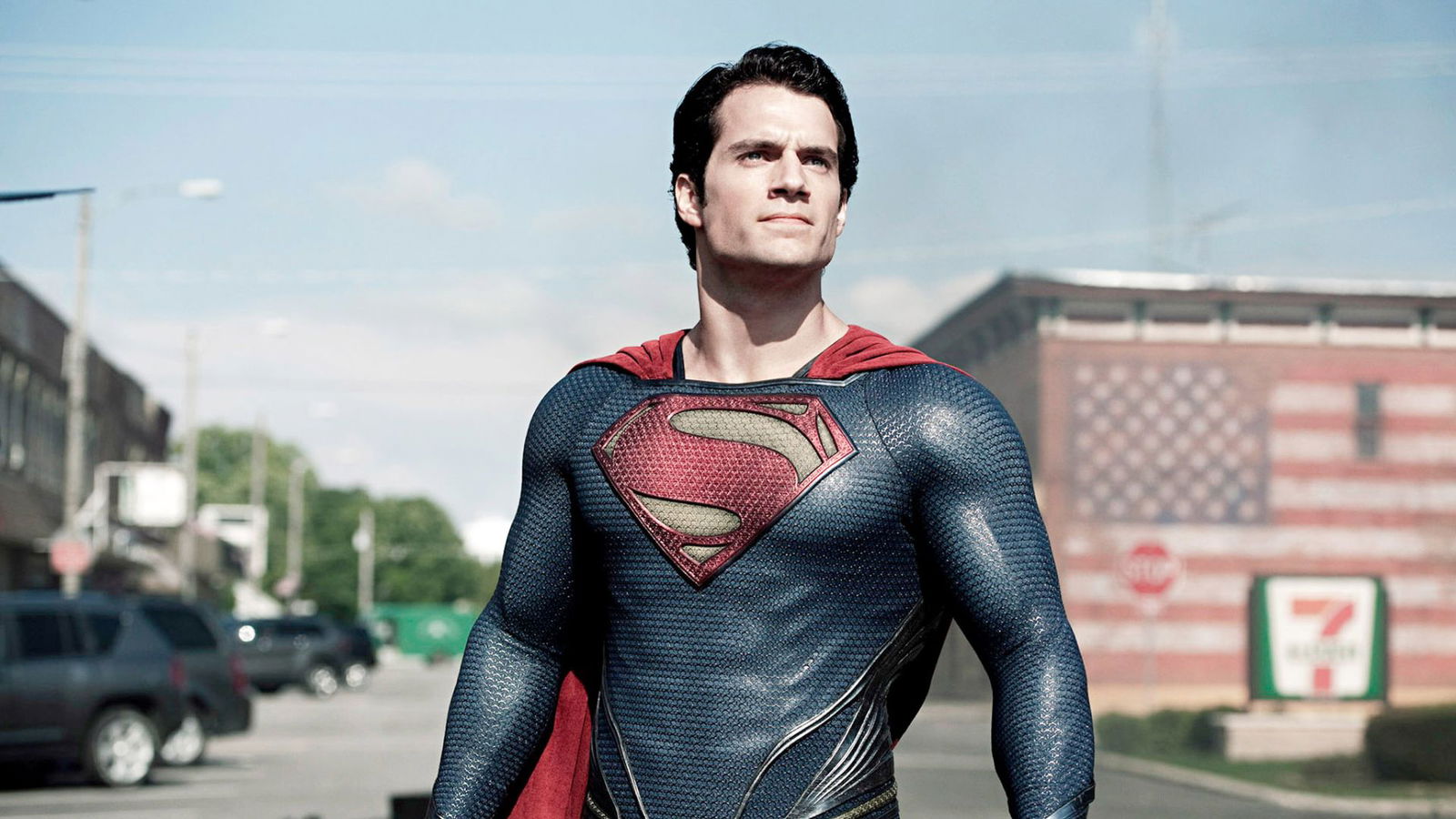 A superhero in a textured blue suit and red cape stands confidently on a city street in daylight, looking toward the horizon.