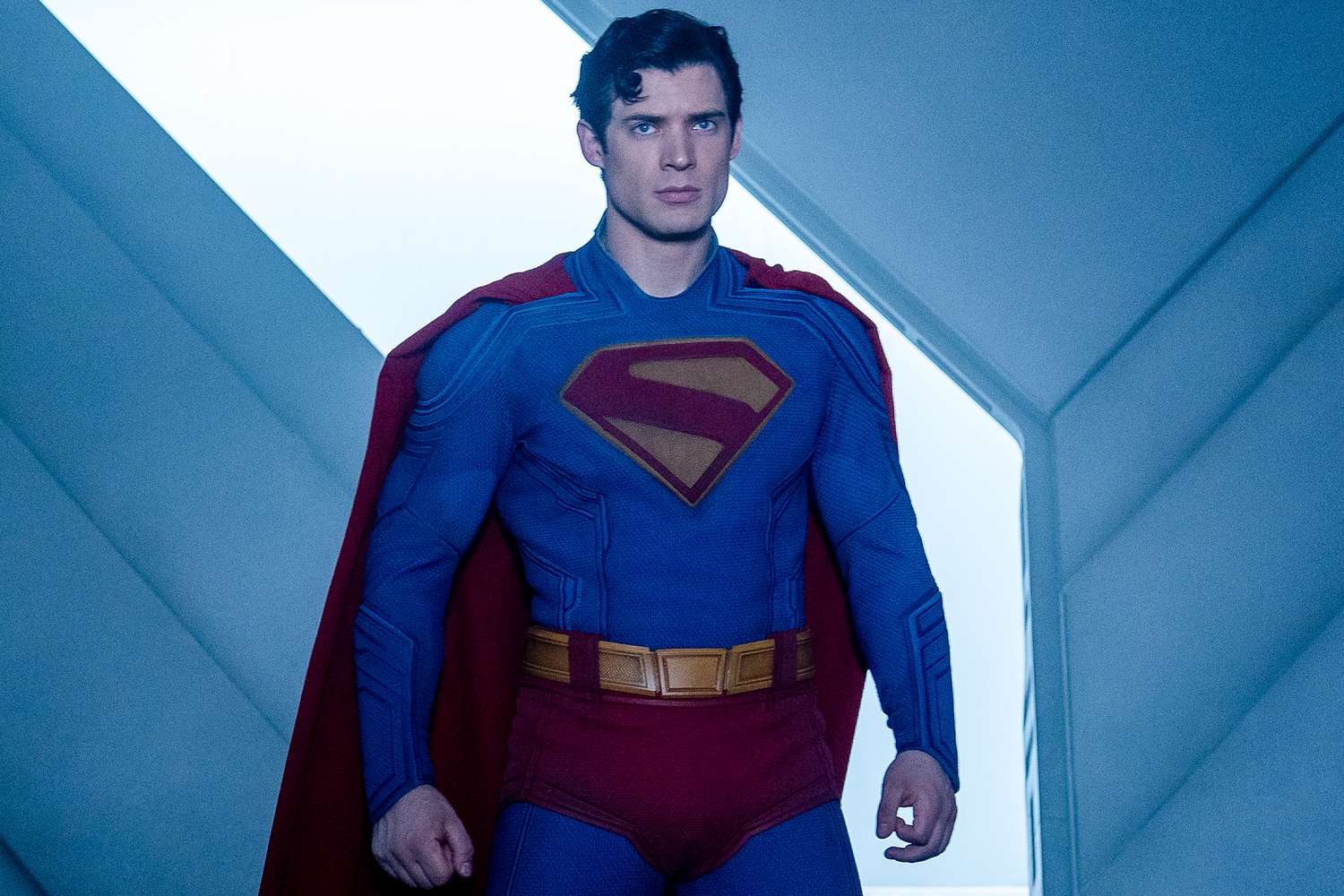 A superhero in a bright blue suit and red cape walks through a futuristic corridor, wearing a classic red-and-yellow chest emblem and a determined expression. The strongest of the kryptonians