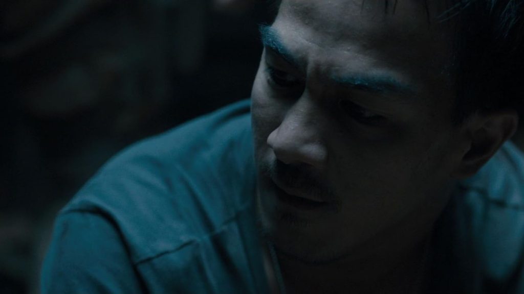 Joe Taslim looks down with a weary, conflicted expression in a dimly lit blue-toned scene. The close-up emphasizes the emotional burden carried by his character, with shadows and sweat highlighting the intensity of the moment.
