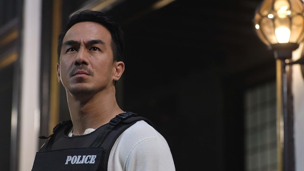 Joe Taslim stands outdoors at night wearing a police tactical vest over a white shirt. He looks into the distance with a focused, determined expression as warm street lighting illuminates the scene behind him.