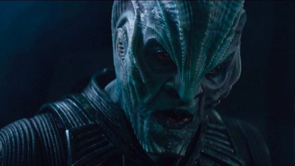 oe Taslim appears in heavy alien makeup and futuristic armor, his face marked by ridged prosthetics and deep shadows. Bathed in cold blue lighting, he stares intensely off-screen in a tense science-fiction setting.