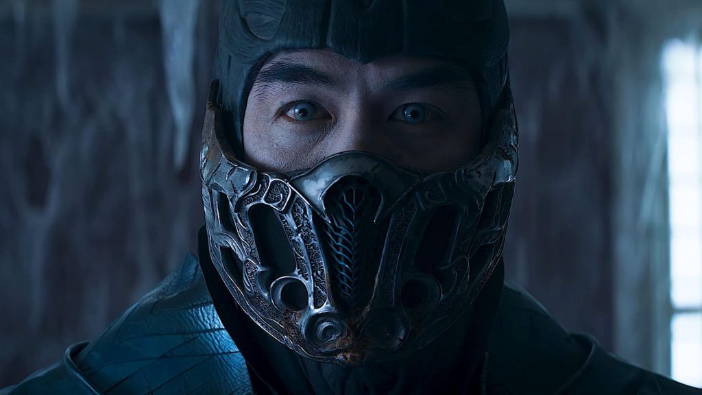Joe Taslim as Sub-Zero stares ahead with icy blue eyes from behind an ornate metallic mask. Framed in a cold, blue-lit environment, the close-up emphasizes the character's intimidating presence and supernatural power.