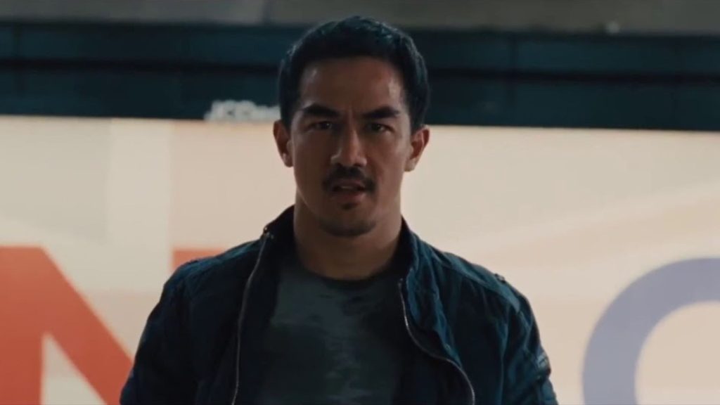 Joe Taslim stands facing forward with a determined expression, wearing a dark jacket over a gray shirt. The scene is lit by bright urban lighting, emphasizing the tension before an impending confrontation.