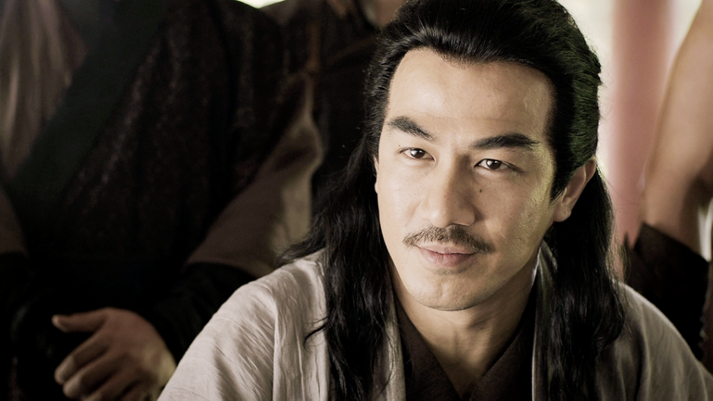 Joe Taslim, dressed in period attire with long flowing hair and a thin mustache, looks confidently toward someone off-camera. Warm indoor lighting and richly detailed costumes evoke the historical setting of the scene.