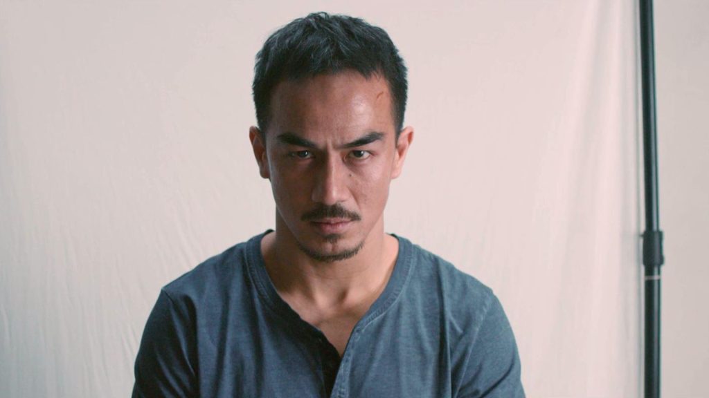 Joe Taslim sits facing the camera with an intense, unblinking stare. Wearing a dark blue henley shirt, he appears bruised and battle-worn, with a small cut on his forehead hinting at recent violence. The minimalist background keeps the focus entirely on his expression.