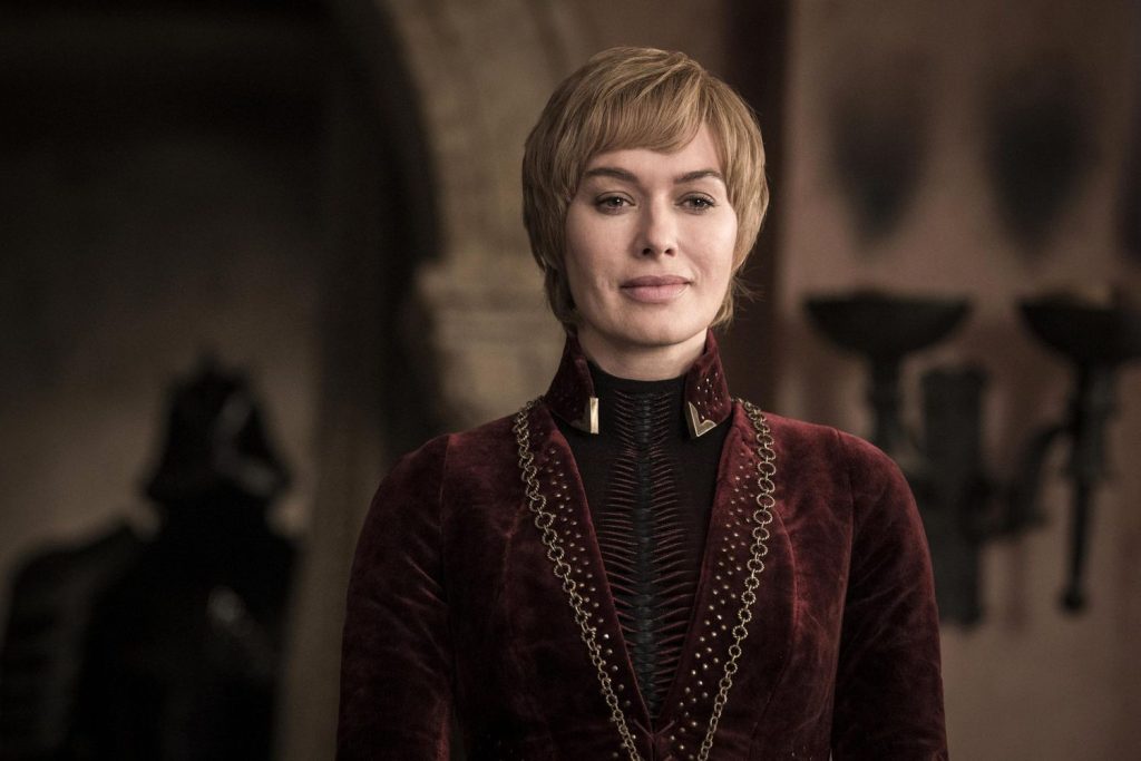 A woman with short blonde hair stands inside a grand hall, wearing an elegant dark red gown with gold detailing. She has a composed expression as the background fades softly out of focus.