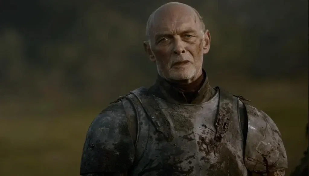 A bald man wearing battered steel armor stands outdoors with a stern expression. His armor is stained from battle, and a blurred landscape stretches behind him.