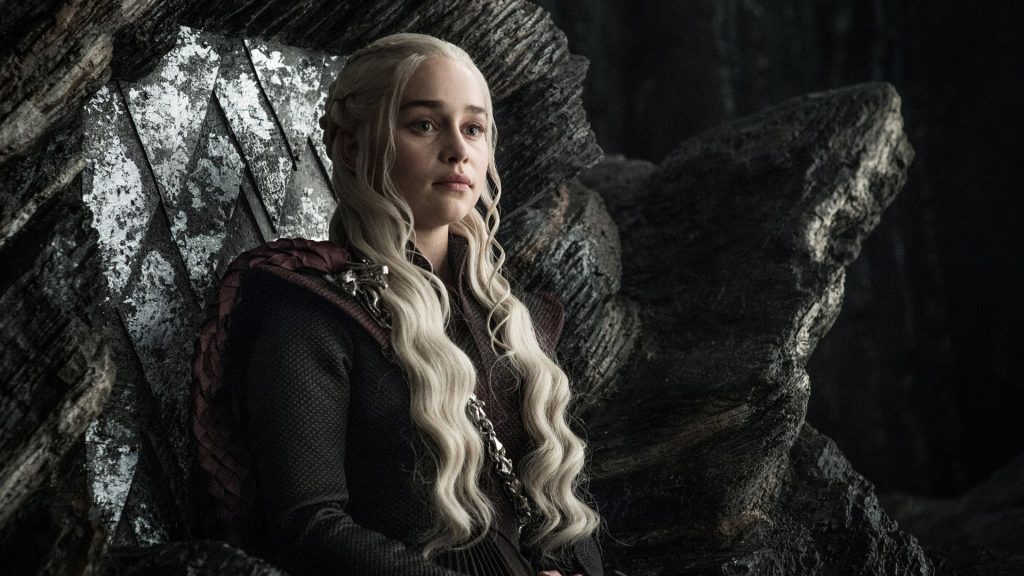 A young woman with long platinum-blonde hair sits on a dark, carved throne, wearing a black outfit with dragon-inspired details. She gazes forward with a calm, determined expression in a dimly lit hall. She is Daenerys, belonging to the same family as Rhaenyra from House of the Dragon