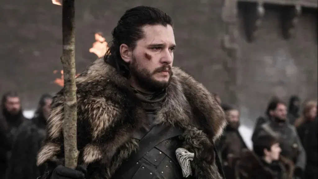 A man with dark hair and a beard stands in a snowy courtyard wearing black armor and a heavy fur cloak. A fresh wound marks his cheek as he grips a torch, with soldiers gathered in the background.
