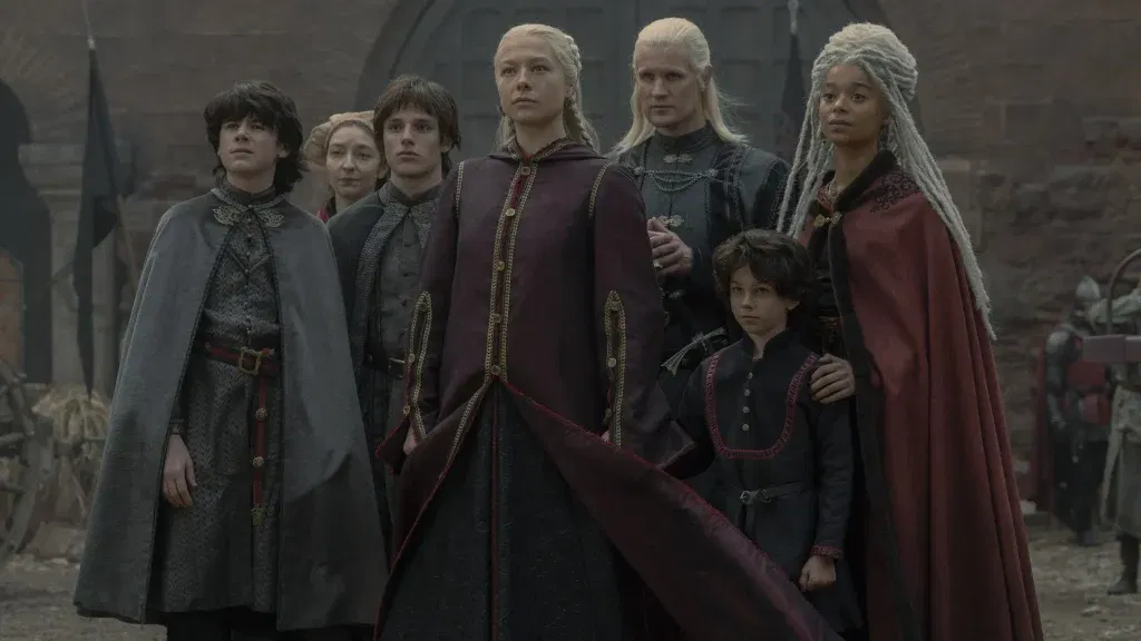 A group of Targaryens and Velaryons stand together in a castle courtyard, with Rhaenyra Targaryen at the center flanked by Daemon Targaryen, their children, and Rhaena Targaryen. Dressed in dark red and black attire, they look ahead with determination as Team Black prepares for the coming conflict.