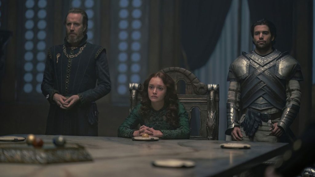 Three members of Team Green stand around the small council table in a dimly lit chamber. Alicent Hightower sits at the center while Ser Criston Cole and another Green supporter stand beside her, discussing strategy as the Dance of the Dragons looms.