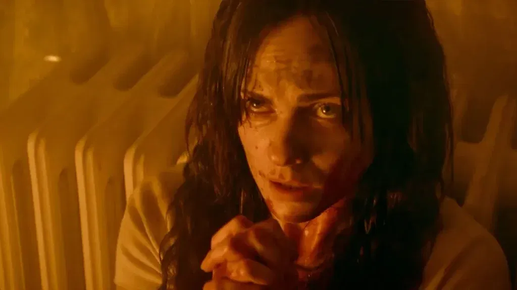 A distressed woman in The Voices of Our Mother with wet, dishevelled hair crouches beside a radiator in a dimly lit room, her face marked with dirt and fear as she stares upward in terror.