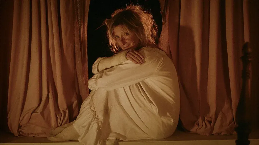 A woman in a white nightgown sits curled up on a window ledge between heavy curtains, gazing forward with an eerie smile in warm, low lighting.