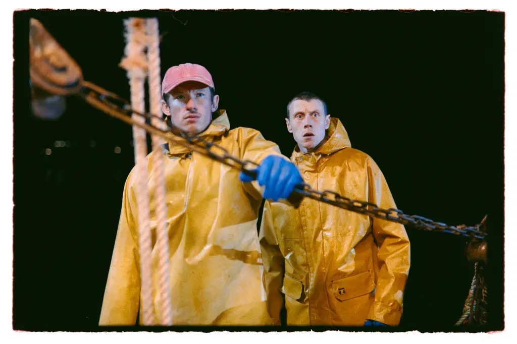 Two fishermen in yellow waterproof gear stand aboard a fishing vessel at night, staring ahead with alarm as they haul heavy chains from the dark sea.