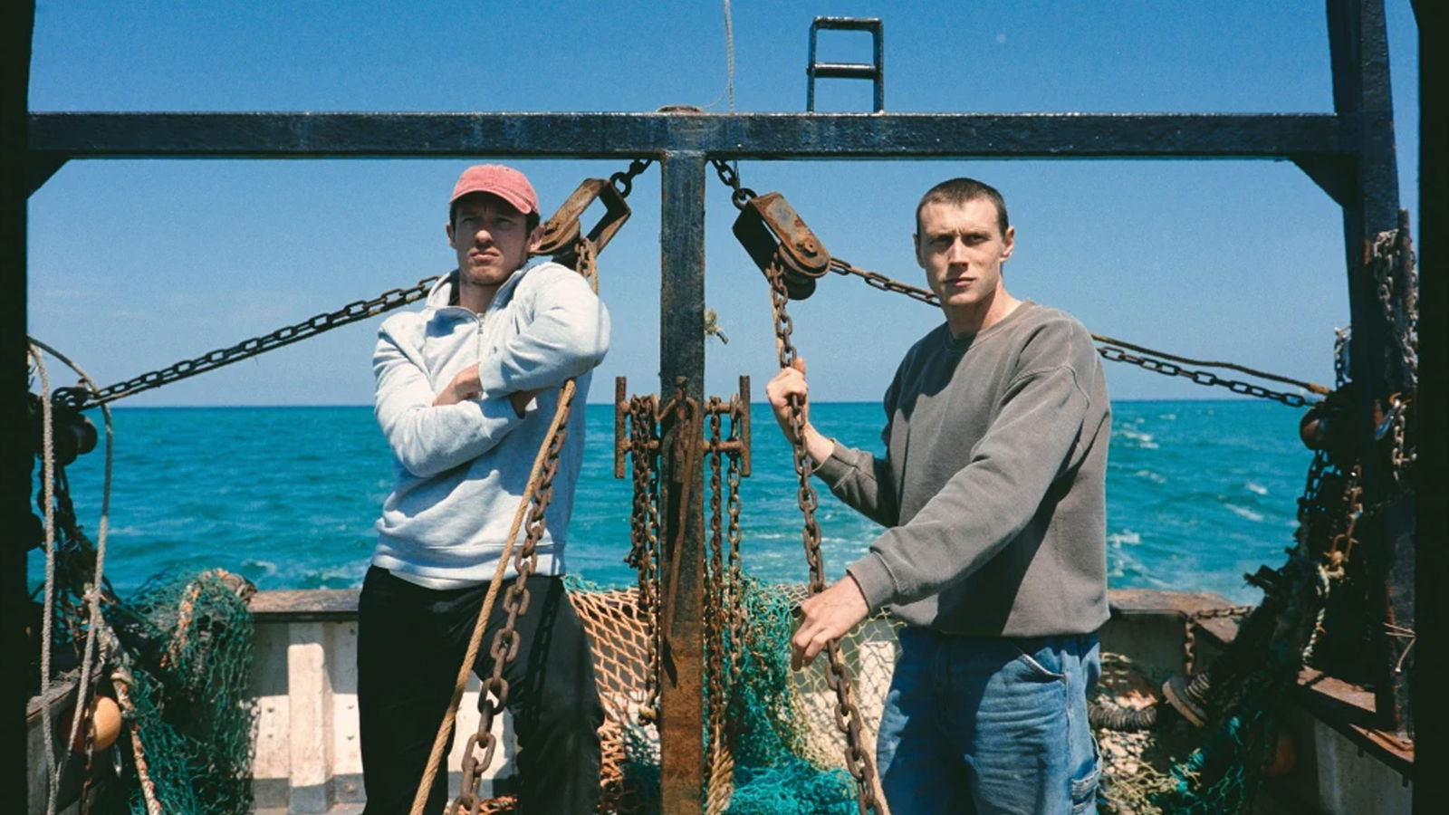Nick and Liam pose aboard the Rose of Nevada during daylight, surrounded by fishing nets, chains, and open sea as they prepare for a routine fishing trip.