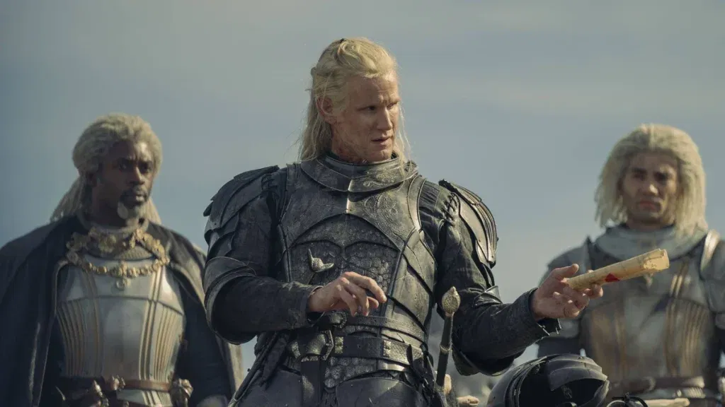 A blond-haired warrior in dark armor studies a sealed scroll while standing outdoors, flanked by two silver-haired companions in armor. The group appears focused on an important message amid a tense military campaign.