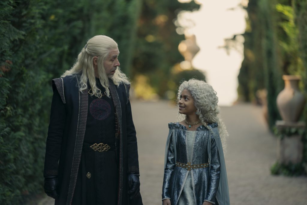 A white-haired nobleman in a dark embroidered robe walks through a garden pathway beside a young girl with curly silver hair wearing a blue and silver dress. The two share a conversation as sunlight filters through the greenery.