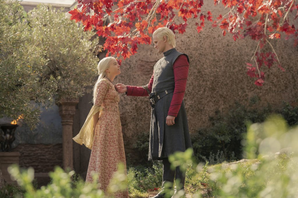 A young silver-haired woman in an ornate patterned gown stands beneath a tree with bright red leaves as a silver-haired man in dark clothing gently touches her chin. The pair share an intimate moment in a sunlit garden courtyard.