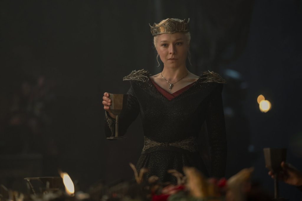 A silver-haired woman wearing a crown and a black gown adorned with dragon-shaped details raises a goblet during a candlelit gathering. She stands solemnly before a banquet table, illuminated by warm torchlight.