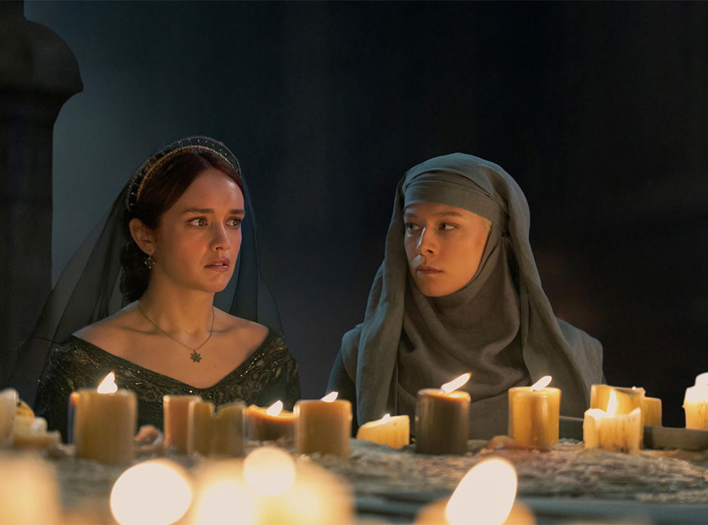 Two women sit beside each other at a candlelit table during a solemn gathering. One wears a green gown and veil, while the other is dressed in a gray hooded robe, both appearing tense and contemplative.