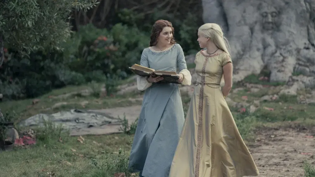Two young noblewomen walk through a garden path while sharing a conversation. One carries an open book, and both smile warmly at each other amid the greenery and stone carvings surrounding them.