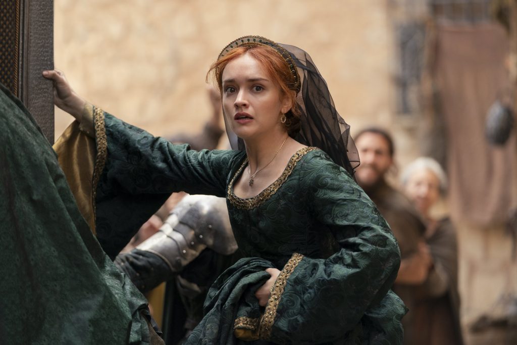 A red-haired woman wearing an ornate dark green gown and black veil looks alarmed as she moves through a crowded street. Townspeople and guards stand behind her, while her expression suggests growing tension and unrest.