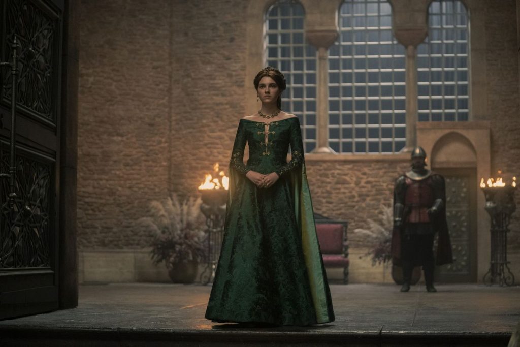 A woman in an elegant dark green gown stands alone in a vast stone hall illuminated by torchlight. A guard watches from the background as she faces forward with a solemn and determined expression.