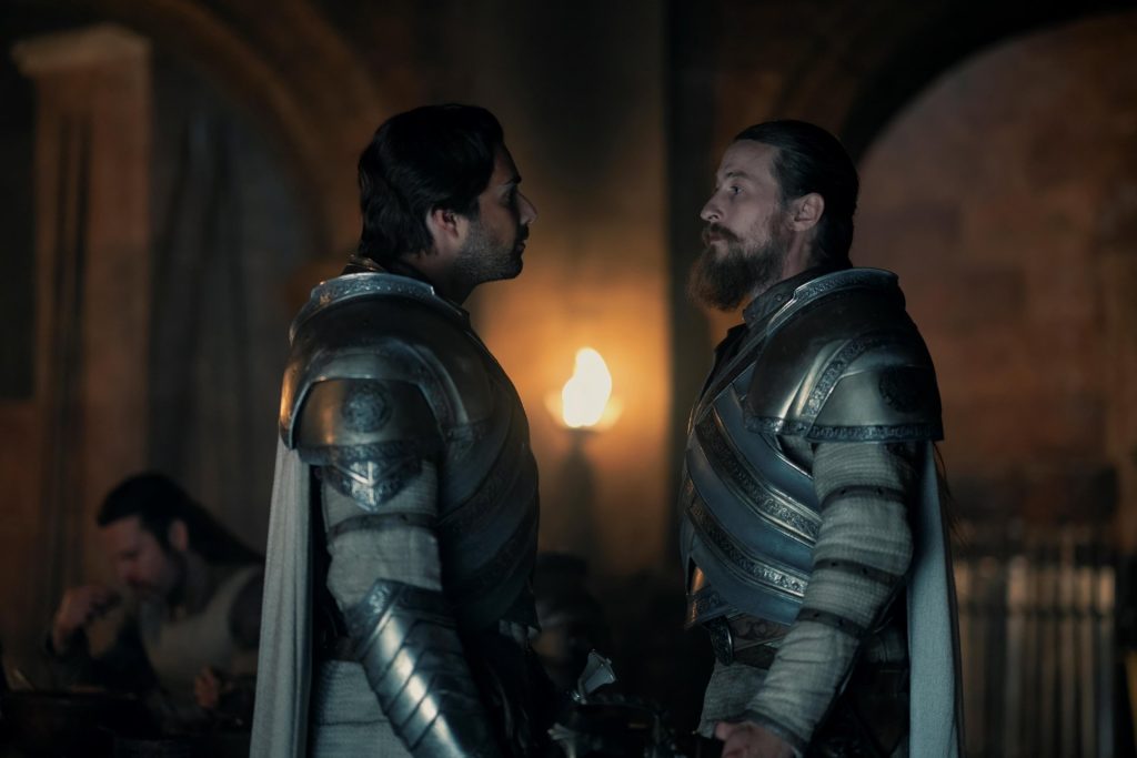 Two armored knights stand face-to-face in a dimly lit hall, their tense expressions suggesting a confrontation. Warm torchlight reflects off their polished armor as onlookers watch from the background.