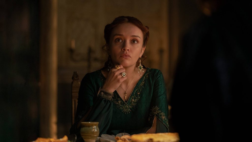 A red-haired woman wearing a dark green embroidered gown sits at a candlelit table, resting her hand near her chin as she listens intently. Her thoughtful expression is illuminated by warm light in a medieval chamber.