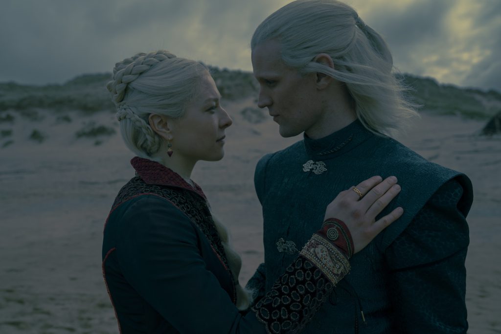 A silver-haired woman and a silver-haired man stand face-to-face on a windswept beach, gazing intently at one another. The woman rests a hand on his chest as storm clouds gather in the distance, creating an intimate and emotionally charged atmosphere.