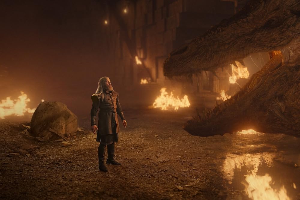 A silver-haired man stands amid flames inside a cavernous dragonpit as the enormous head of a dragon looms before him. Firelight reflects across the scorched ground, creating a tense and dangerous atmosphere.