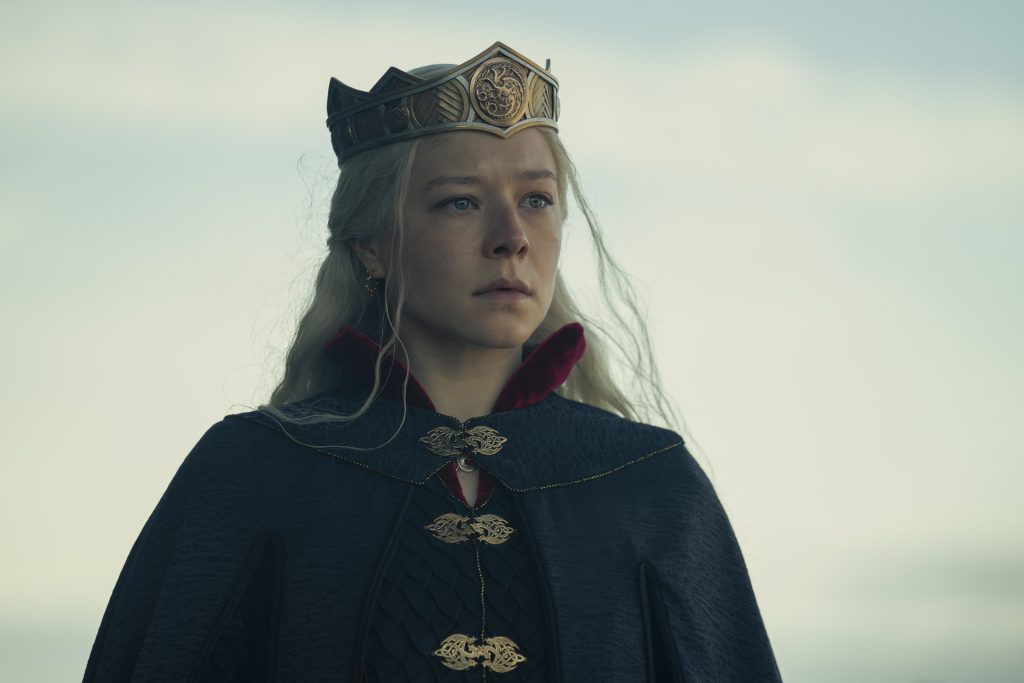A silver-haired woman wearing a bronze crown and a dark cloak stands against a pale sky, her expression solemn and resolute. Fine details on her royal attire catch the light as she gazes into the distance.