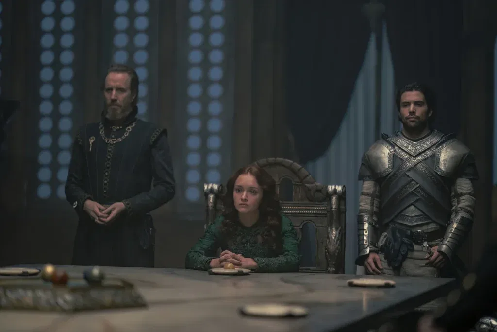Three figures stand around a large council table in a dimly lit chamber. A young woman in a green gown sits at the head of the table while two men, one in dark court attire and the other in armor, stand beside her with solemn expressions.
