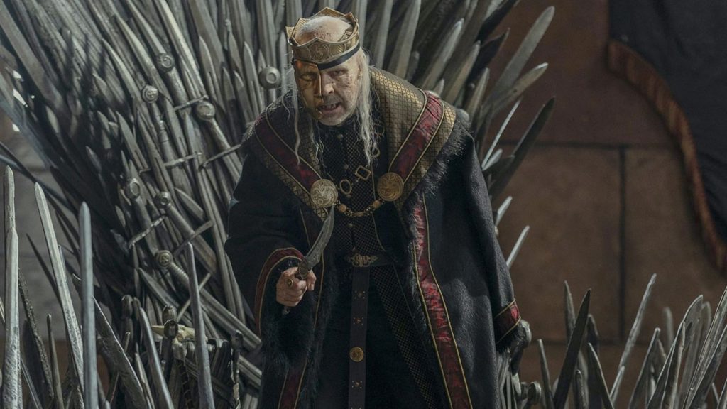 An elderly silver-haired king wearing a crown and dark royal robes stands before the Iron Throne, gripping a dagger as he addresses the court. The towering swords of the throne loom behind him, emphasizing the weight of his authority and frailty.