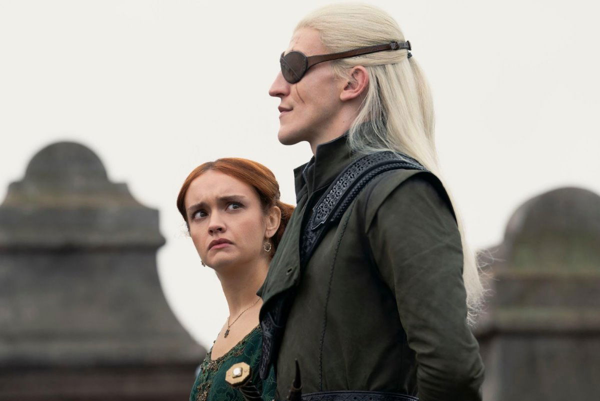 A red-haired woman in a dark green gown stands beside a silver-haired man wearing a black eyepatch and dark leather attire. She looks concernedly at the man.