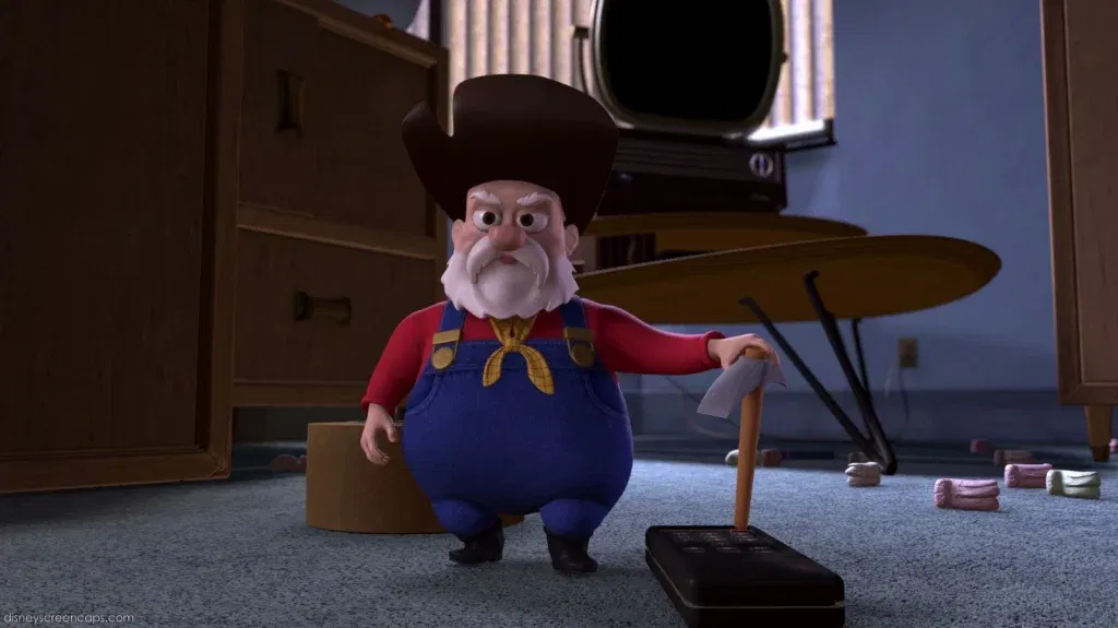 Stinky Pete, the Prospector from Toy Story 2, stands sternly in a room while holding a toy pickaxe beside a television remote.