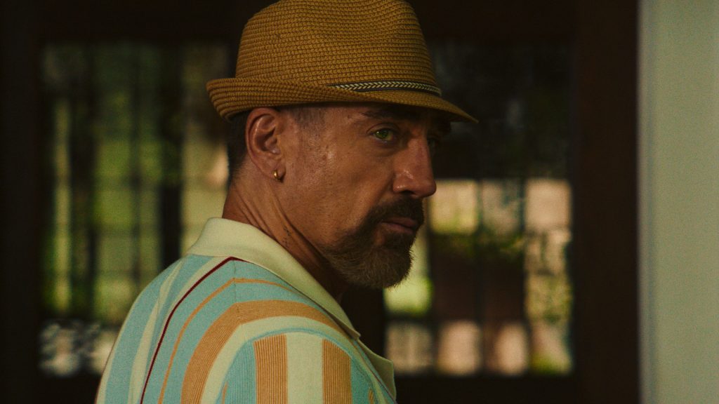 Javier Bardem as Max Cady looks back over his shoulder with a cold, calculating stare while wearing a straw hat and striped polo shirt in Cape Fear Episode 5.