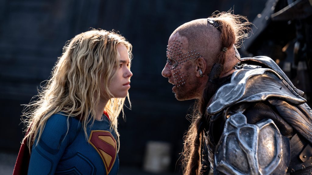 Supergirl confronts Krem of the Yellow Hills in a tense face-to-face standoff, with the armored villain glaring back as the two prepare for battle.