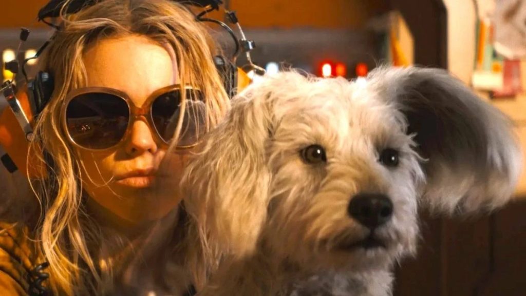 A blonde young woman wearing oversized sunglasses and a head-mounted device stands beside a fluffy white dog, both looking intently ahead inside a warmly lit room.