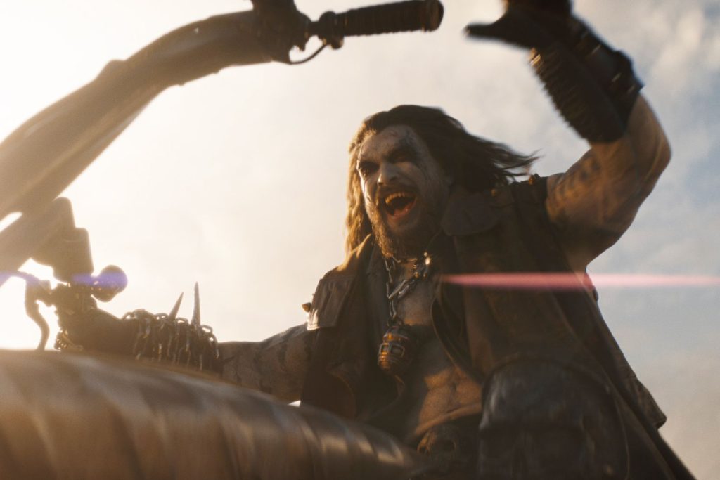A rugged, long-haired man with face paint raises a curved blade while riding a futuristic motorcycle, shouting as he charges into battle.